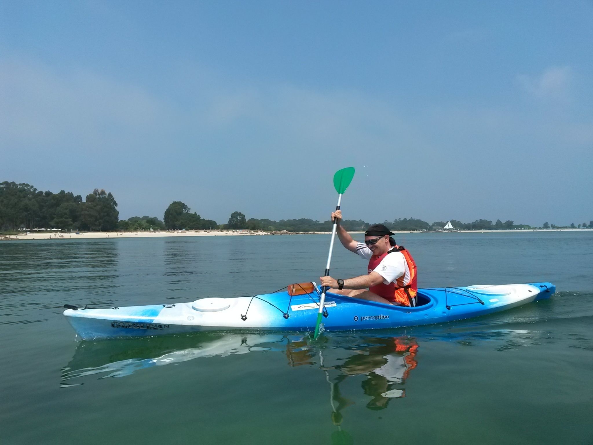 A person kayaking on a calm body of water with a beach and trees in the background.