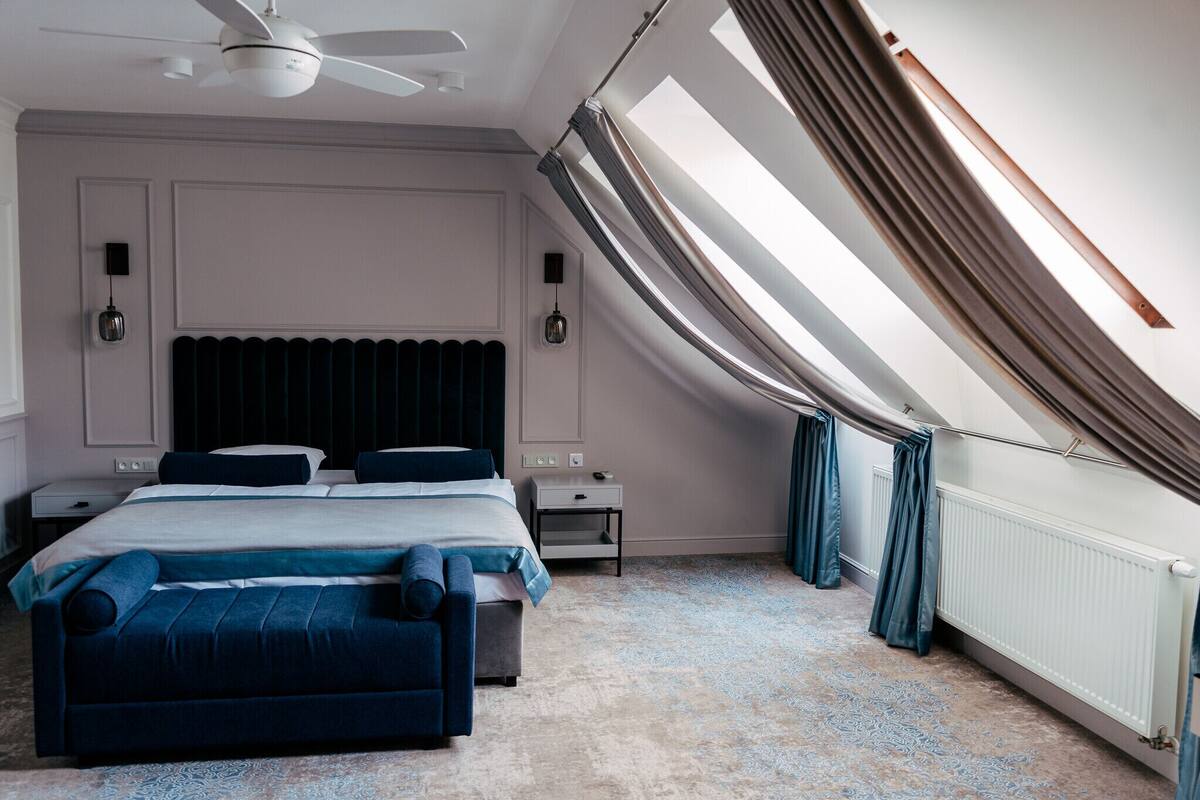 Un dormitorio con cama, mesitas de noche, ventilador de techo, radiador y ventana con cortinas.