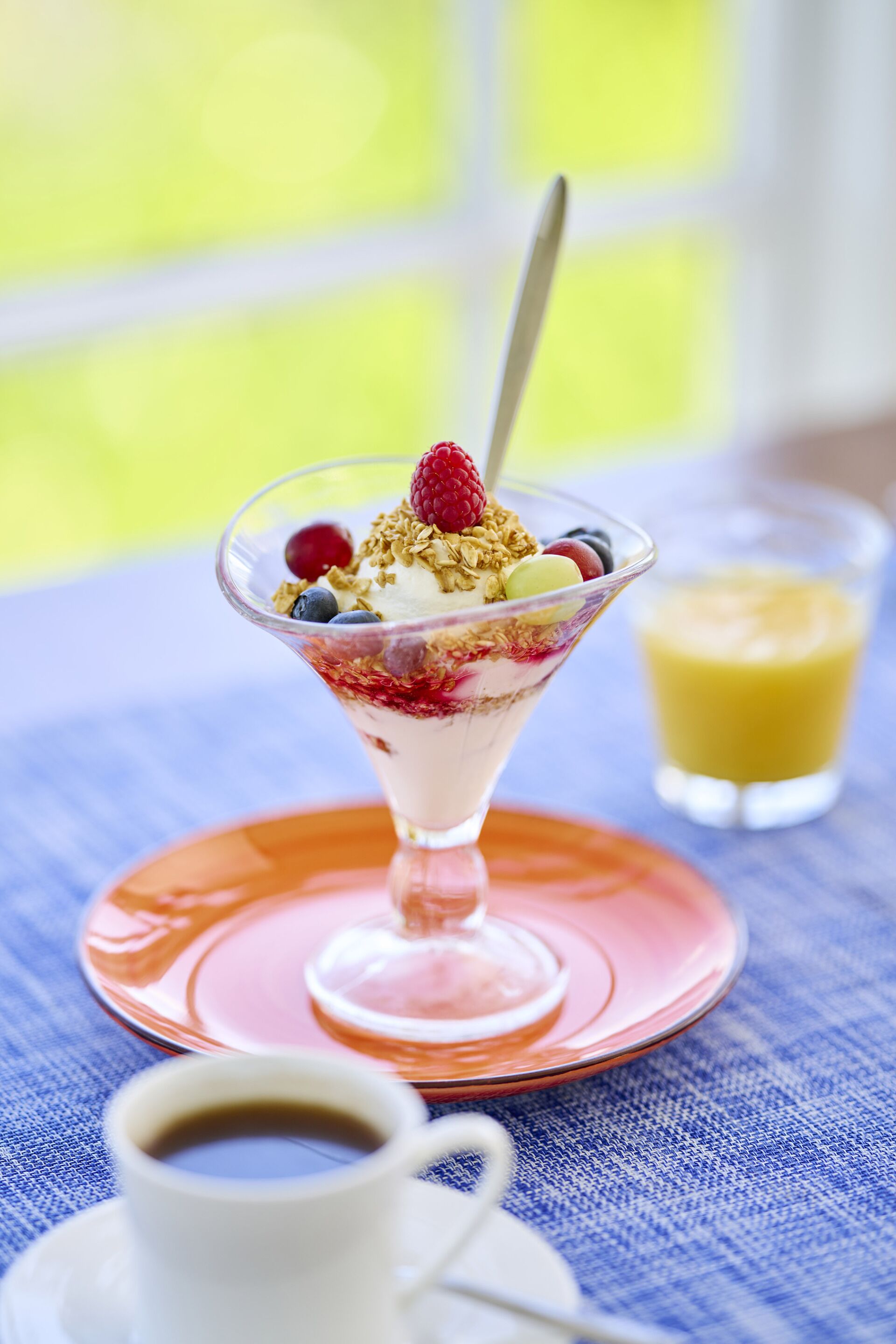 A dessert in a glass bowl on a pink plate, a cup of coffee, and a glass of orange juice on a blue tablecloth.