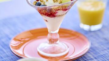 A dessert in a glass bowl on a pink plate, a cup of coffee, and a glass of orange juice on a blue tablecloth.