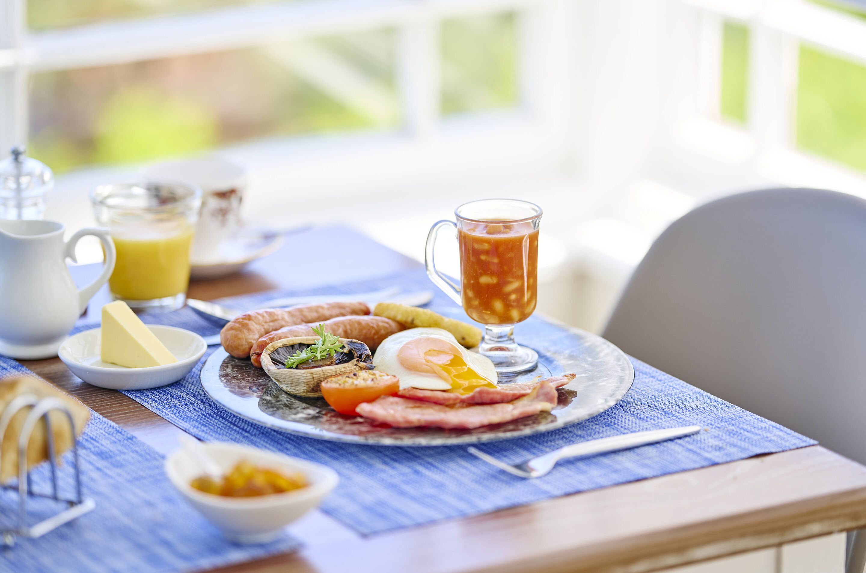 Una mesa de desayuno con un plato de huevos, tocino, tomates y tostadas, un vaso de jugo de naranja y una tetera de té.