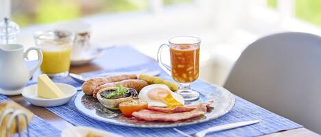 A breakfast table with a plate of eggs, bacon, tomatoes, and toast, a glass of orange juice, and a jug of tea.