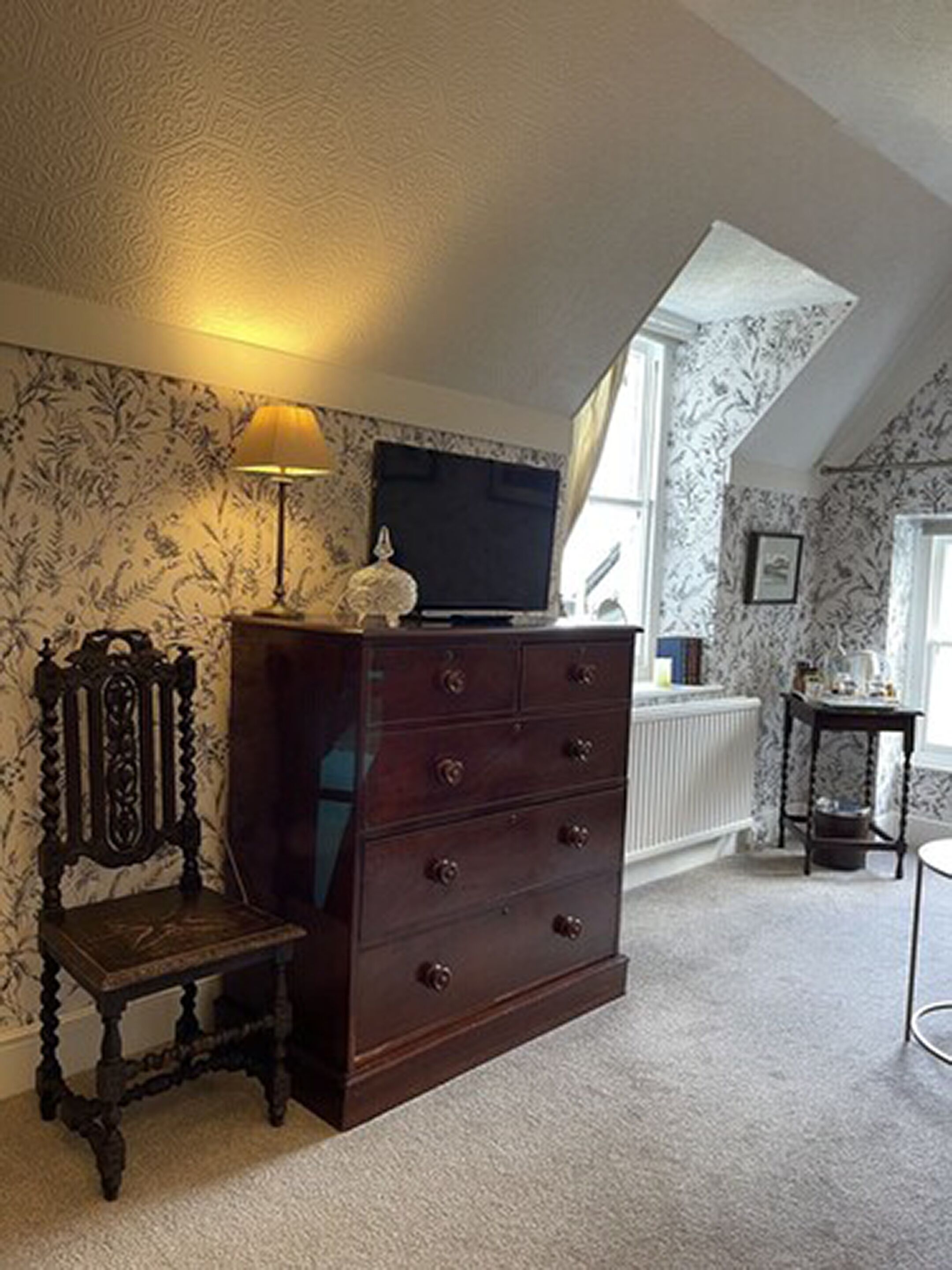 A room with a dark wooden dresser, a lamp, a television on the wall, and patterned wallpaper.