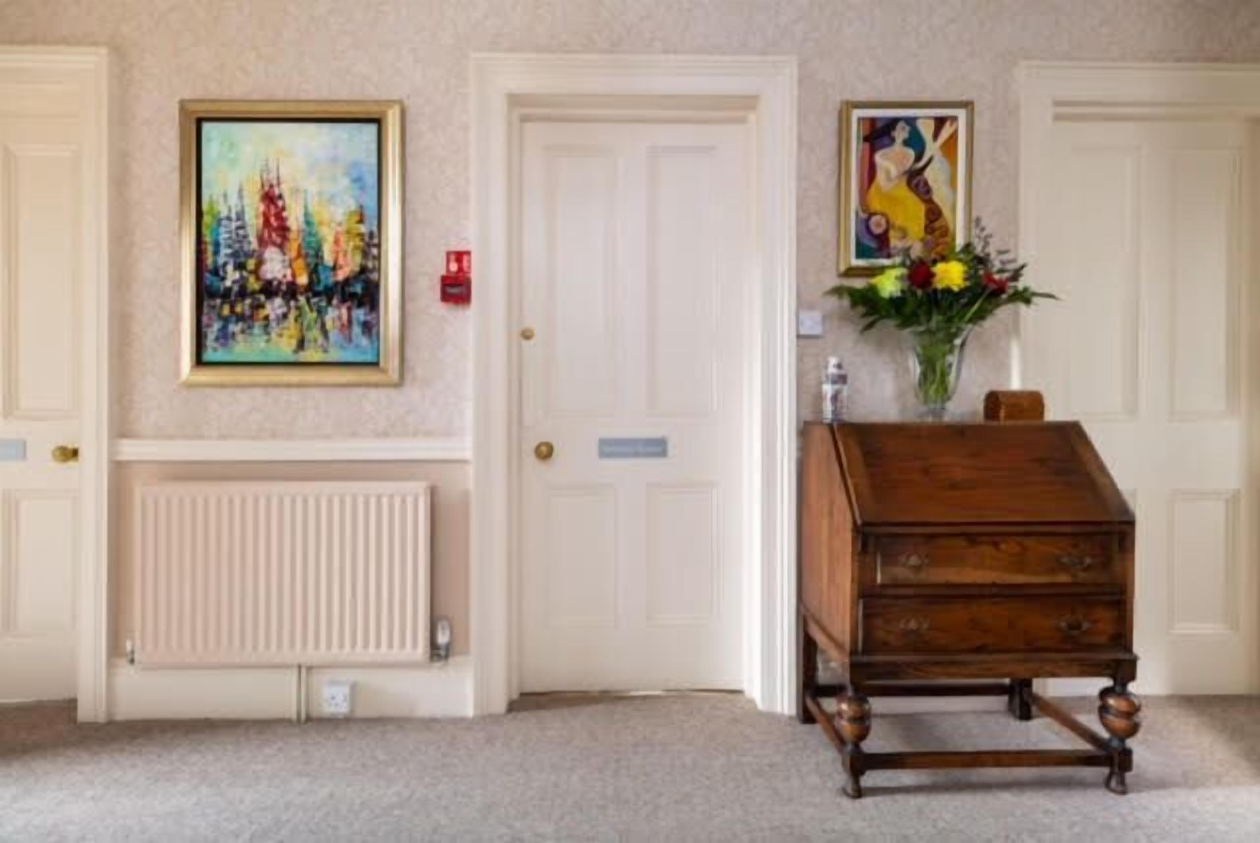 A room with a radiator, a wooden chest of drawers, and two framed paintings on the wall.