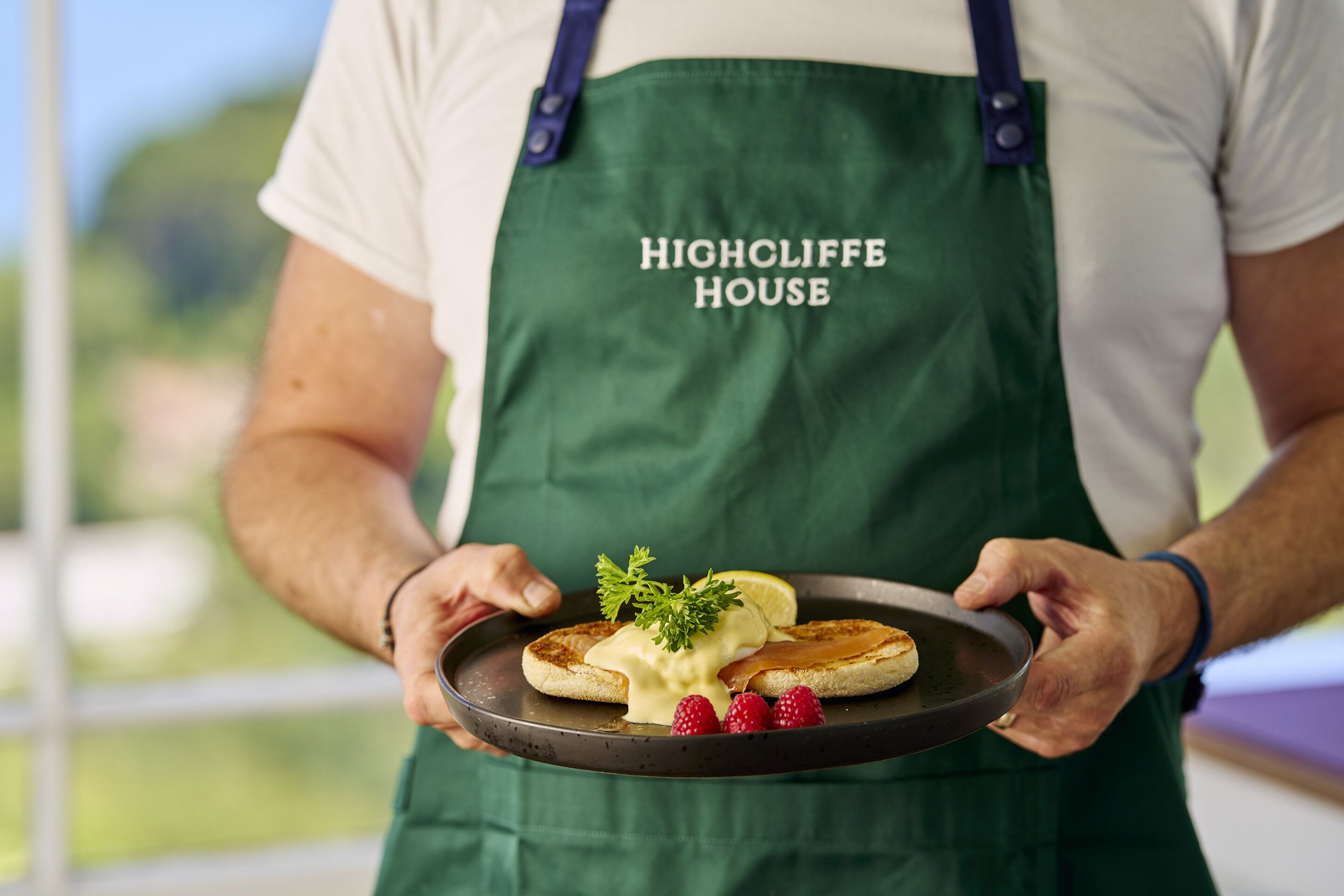 A person holding a plate with a breakfast dish, wearing a green apron with "HIGHOLIFE HOUSE" text.