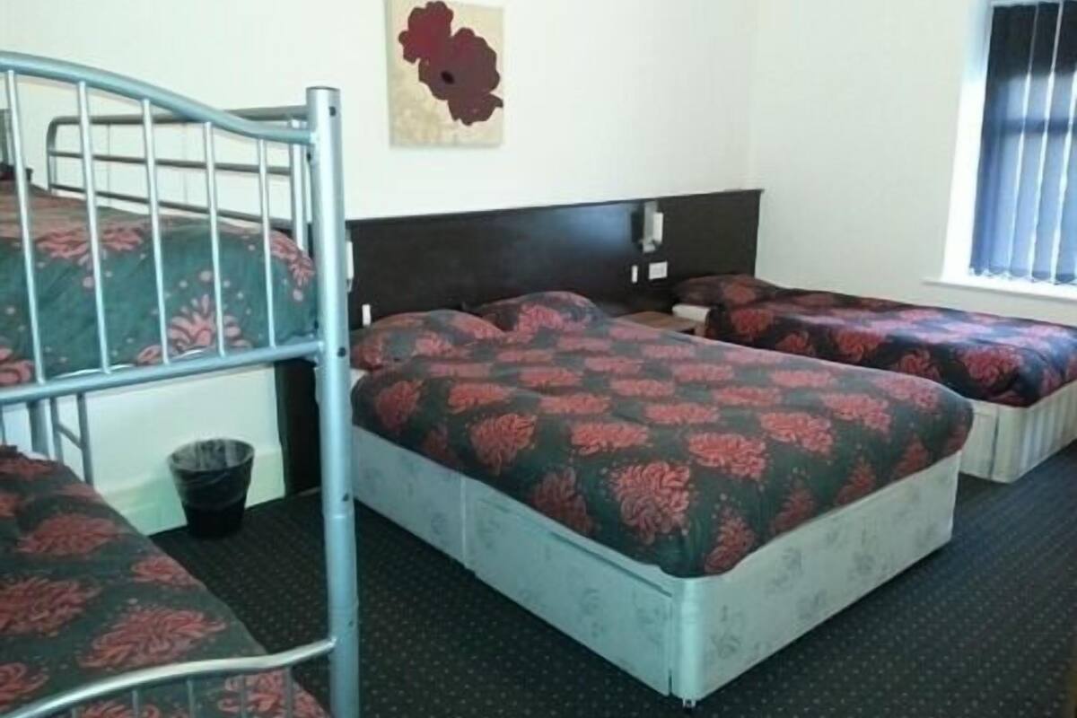 A dormitory room with two beds, a bunk bed, a wall-mounted artwork, and a window with blinds.