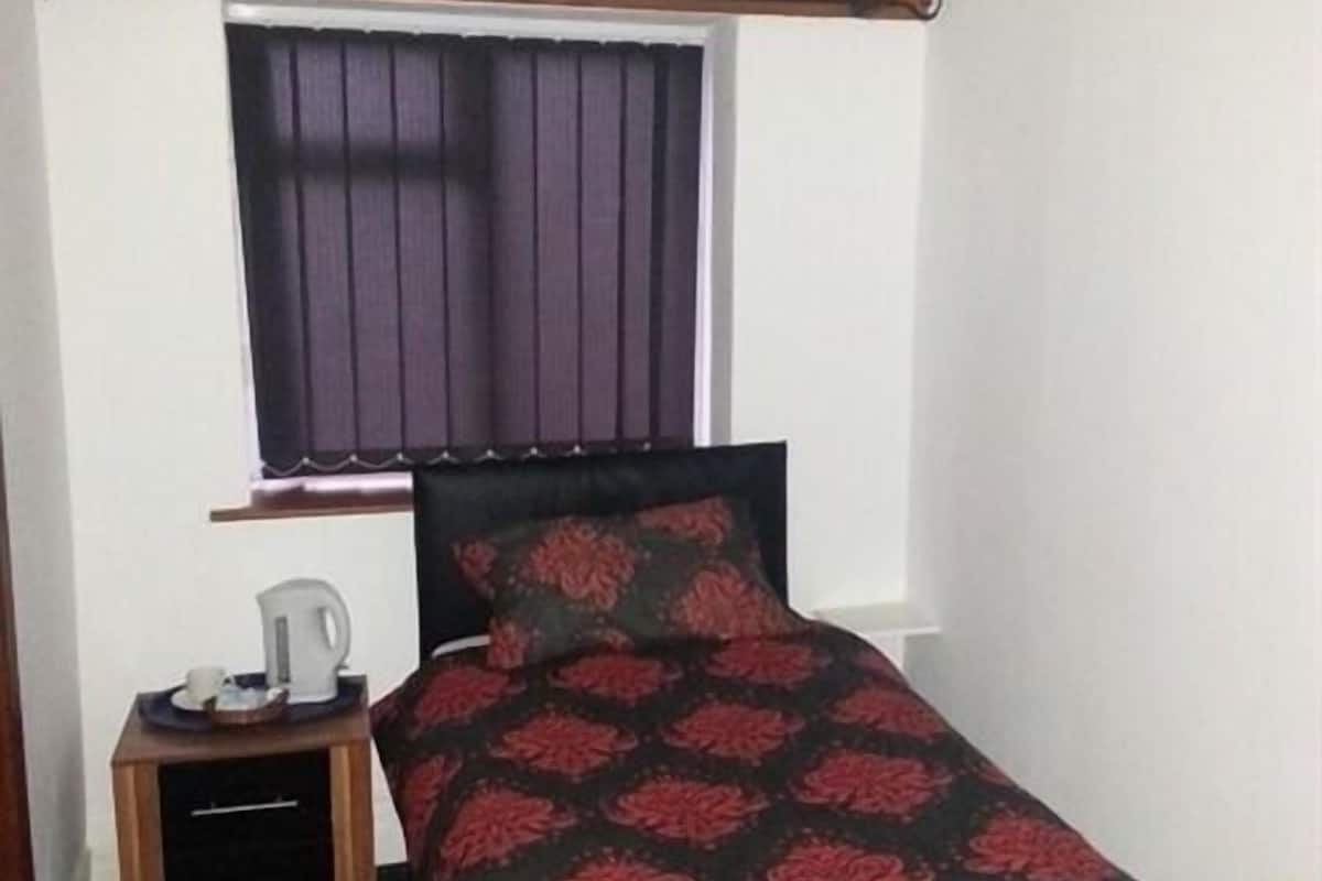 A small, single-bed room with a window, a bedside table with a lamp, and a dark floral bedspread.
