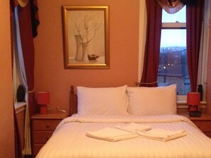 Ein Hotelzimmer mit einem Bett, einem Nachttisch, einer Lampe, einem gerahmten Bild an der Wand und einem Fenster mit Vorhängen.