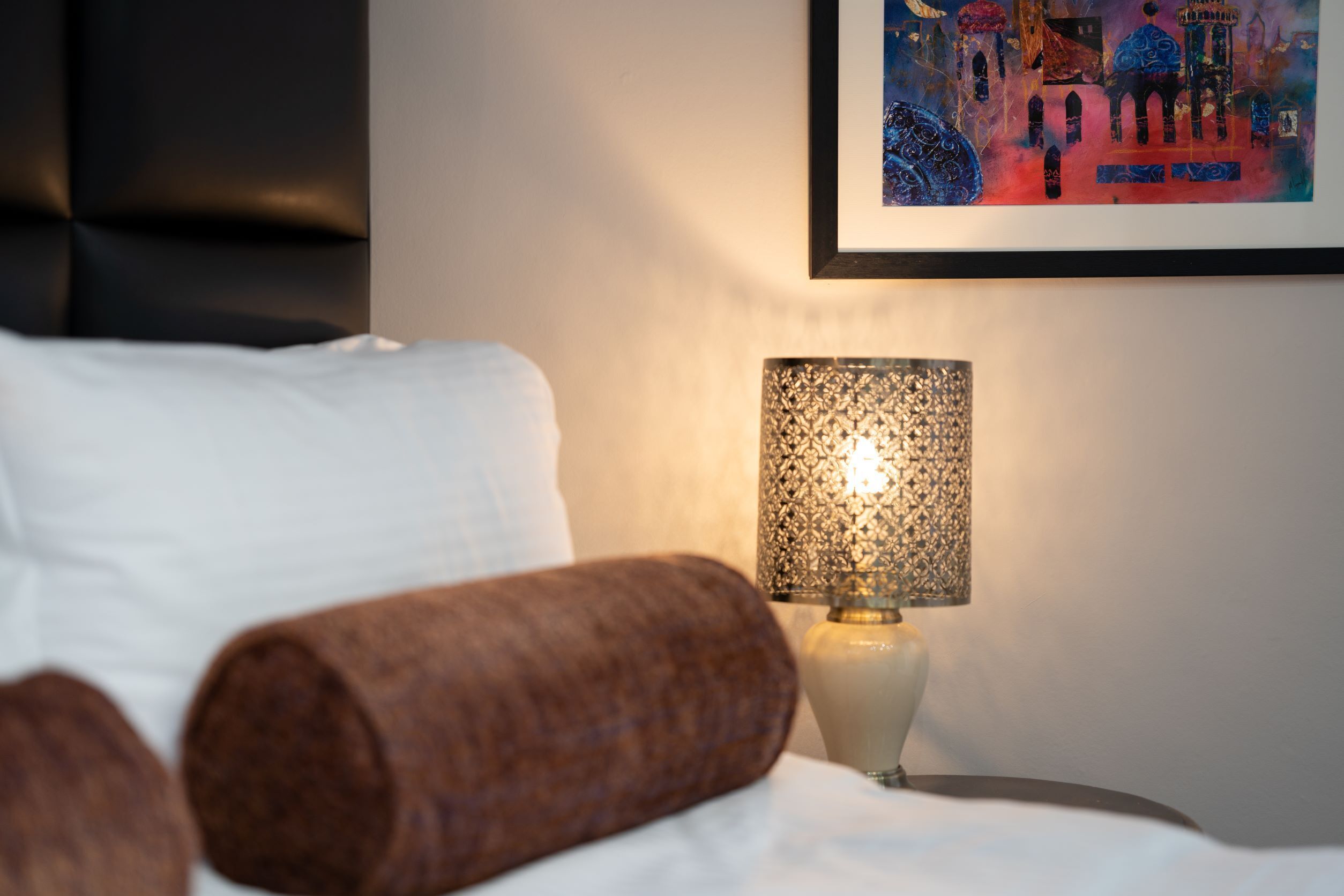 Una cama bien hecha con una colcha blanca y dos almohadas marrones cilíndricas. Una lámpara de pared decorativa con pantalla estampada está colocada sobre una mesita de noche.