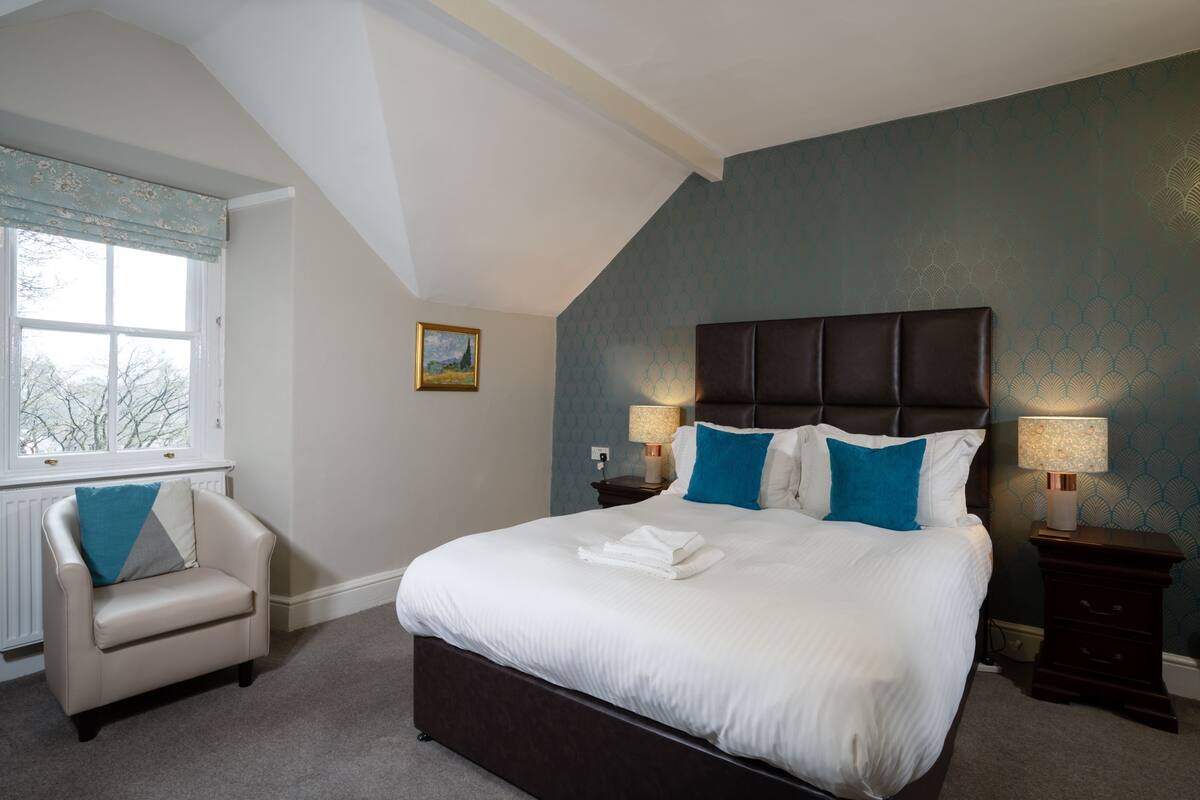 A bedroom with a large bed, two bedside tables with lamps, a chair, and a window with a view of trees.