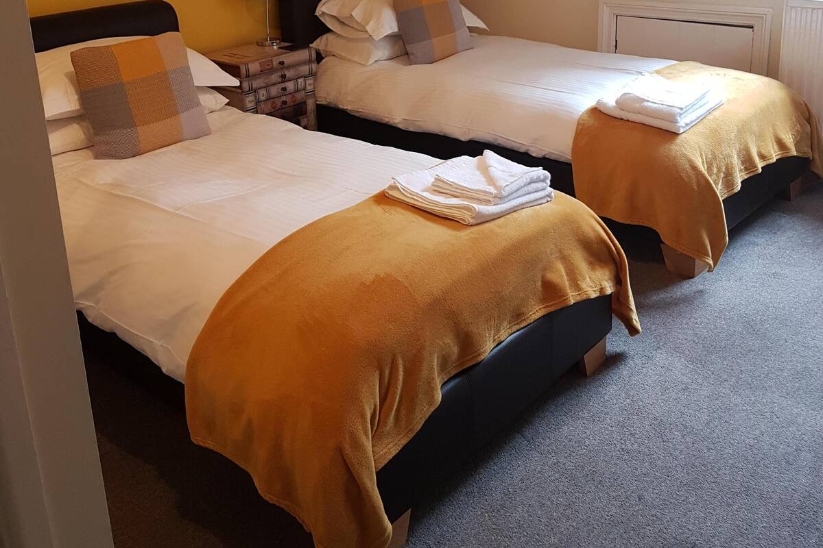A room with two beds, each with a headboard, a nightstand, a lamp, and a window.