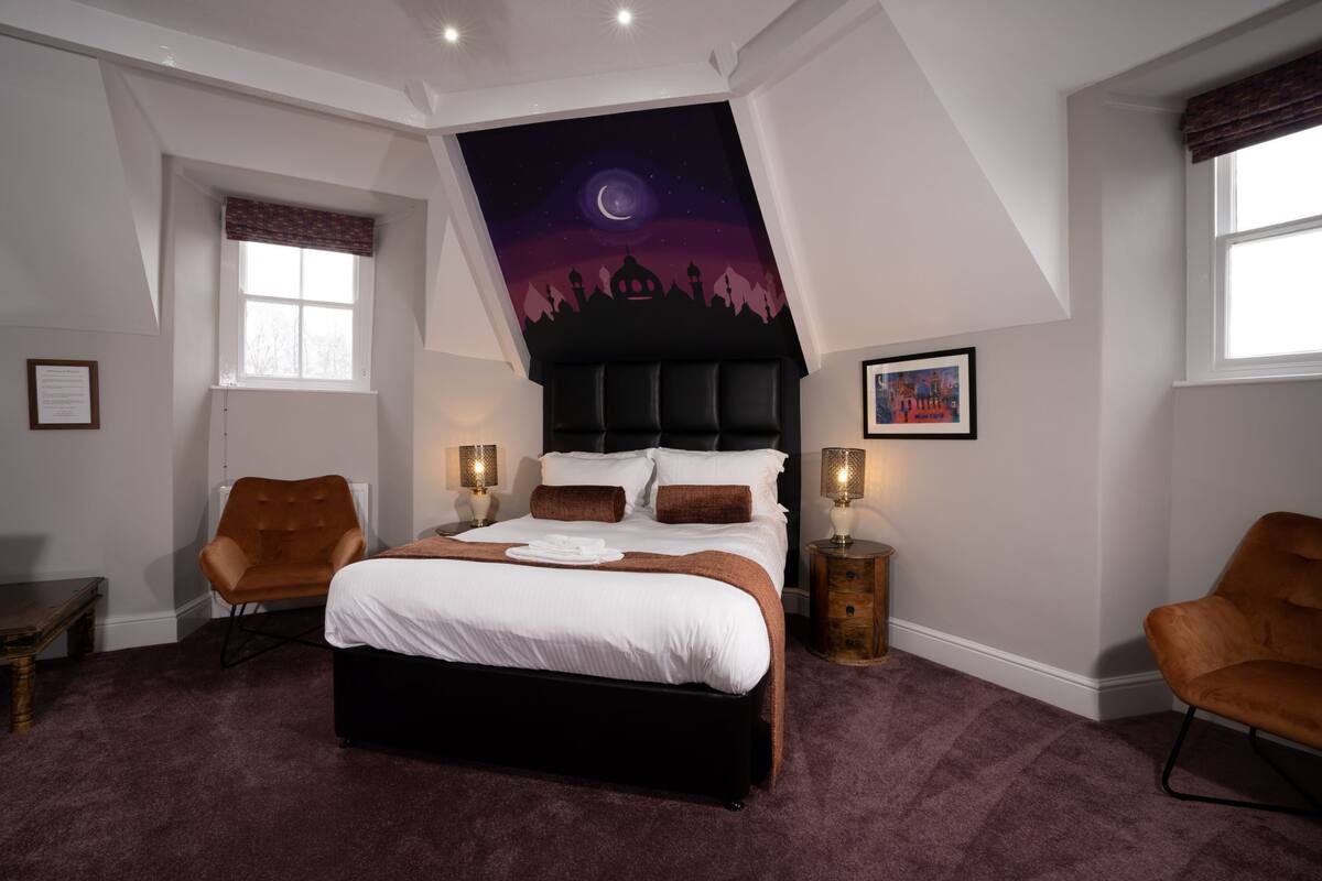 A bedroom with a large bed, two bedside tables, a nightstand, a chair, and a ceiling mural.