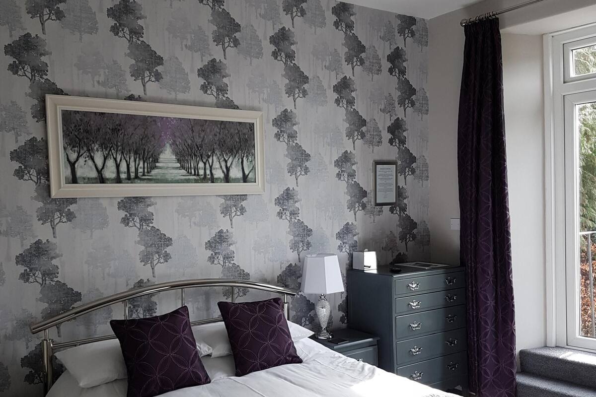 A neatly made bed with a metal frame, a bedside table with a lamp, a dresser, and a window with curtains.