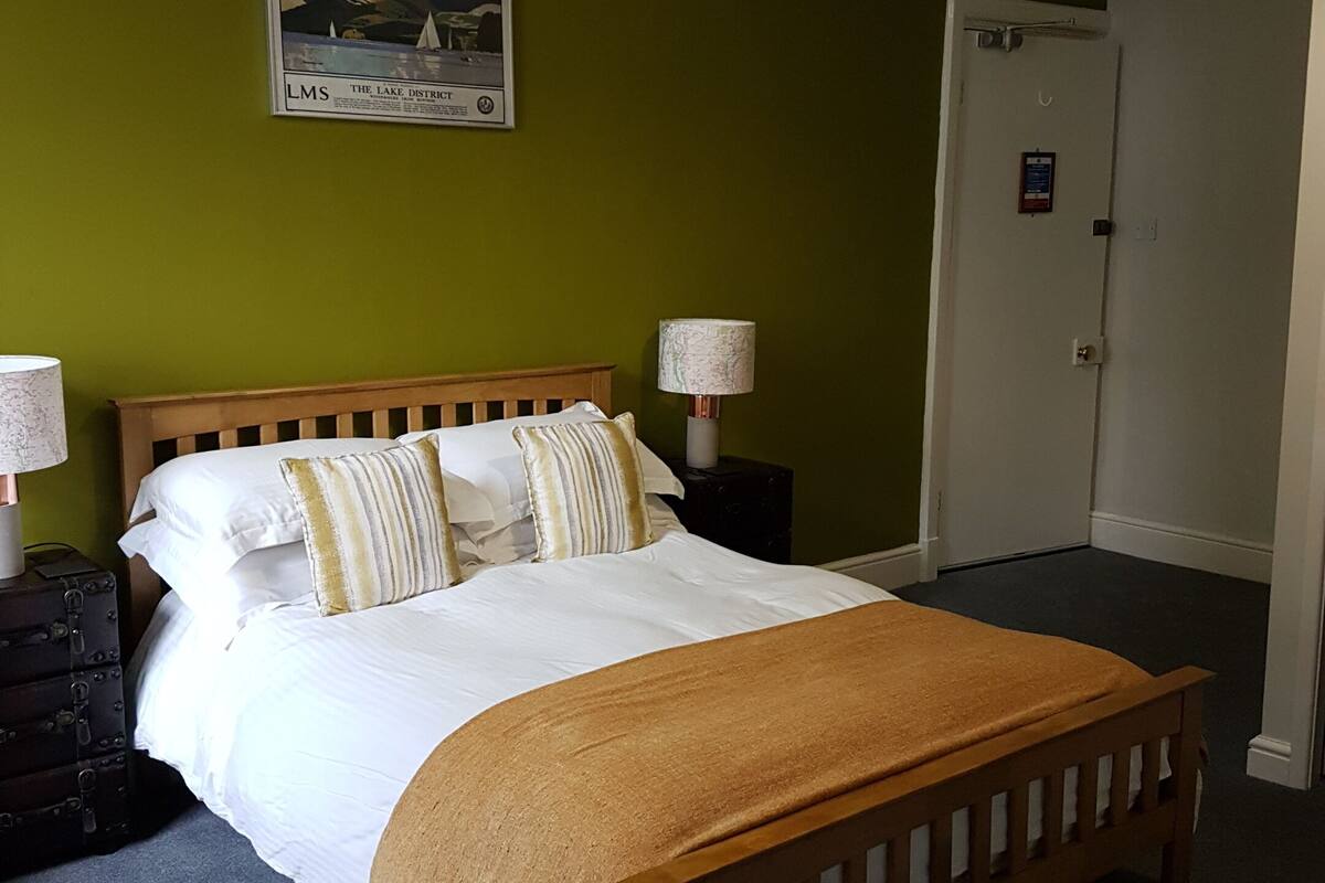 A neatly made bed with a wooden headboard, a brown throw blanket, and striped pillows. There is a framed picture on the wall above the bed, a lamp on a bedside table, and a suitcase on the floor.