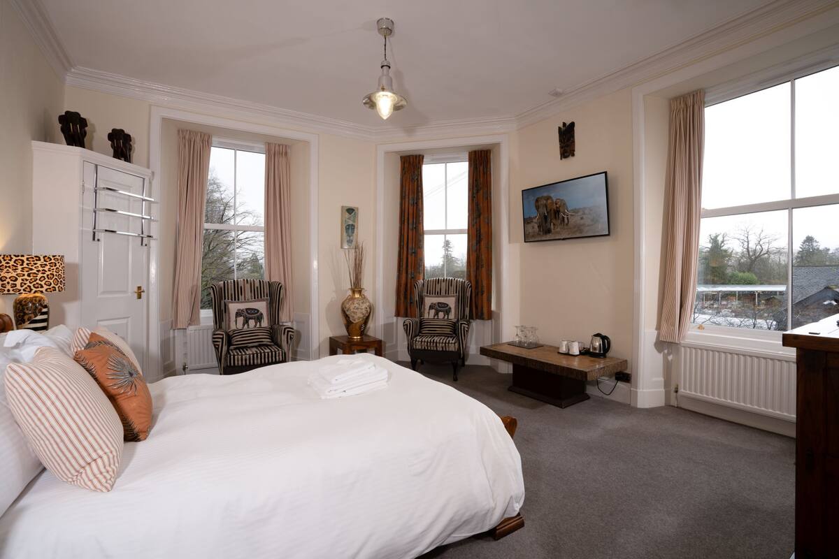 A spacious bedroom with a large bed, a desk, and a chair. There are windows with curtains, a radiator, and a painting on the wall.