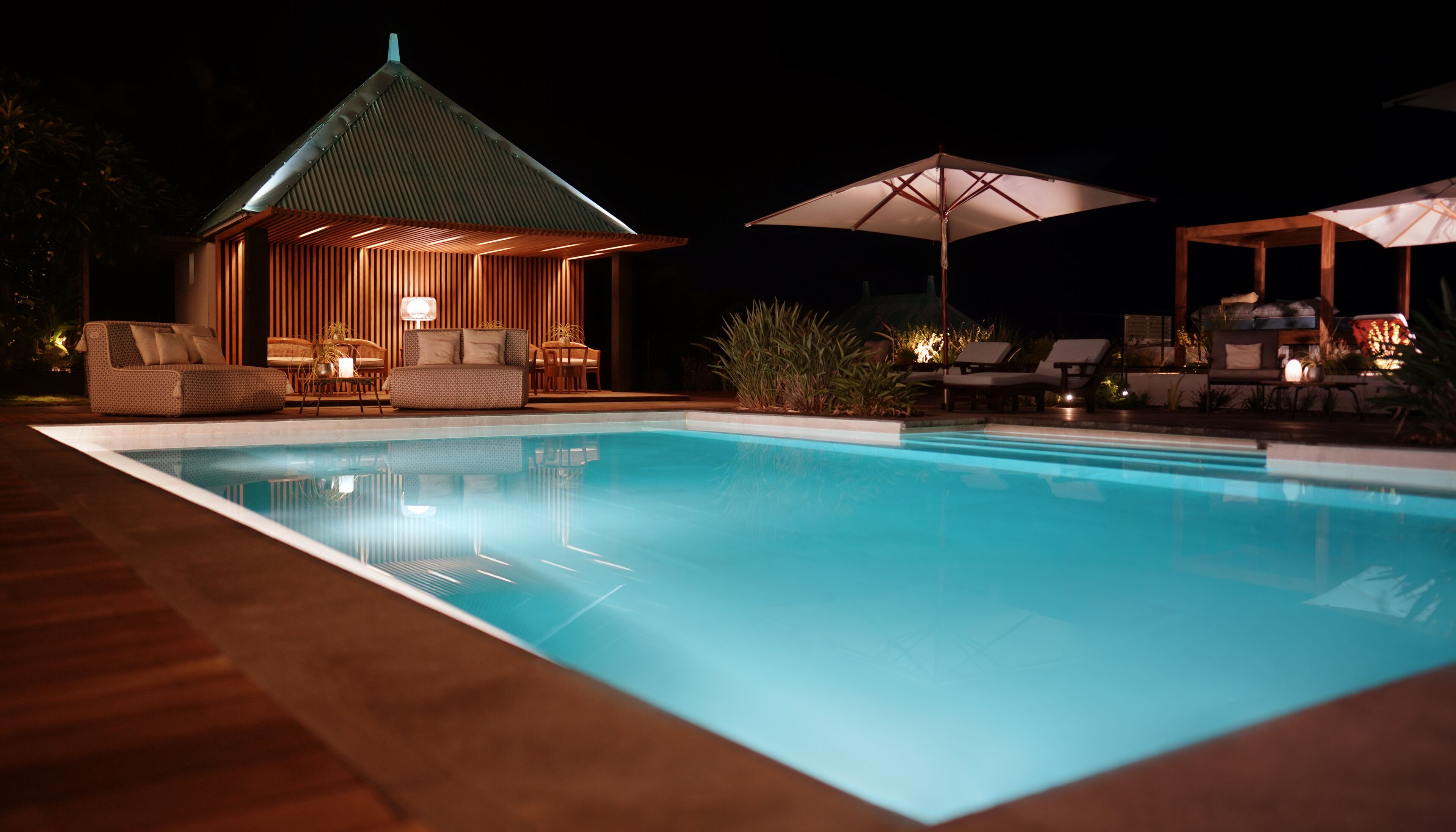A nighttime pool area with a lounge chair and umbrella, a small wooden structure with a sloped roof, and a pool with clear water.