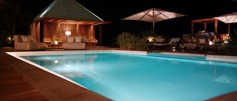 A nighttime pool area with a lounge chair and umbrella, a small wooden structure with a sloped roof, and a pool with clear water.