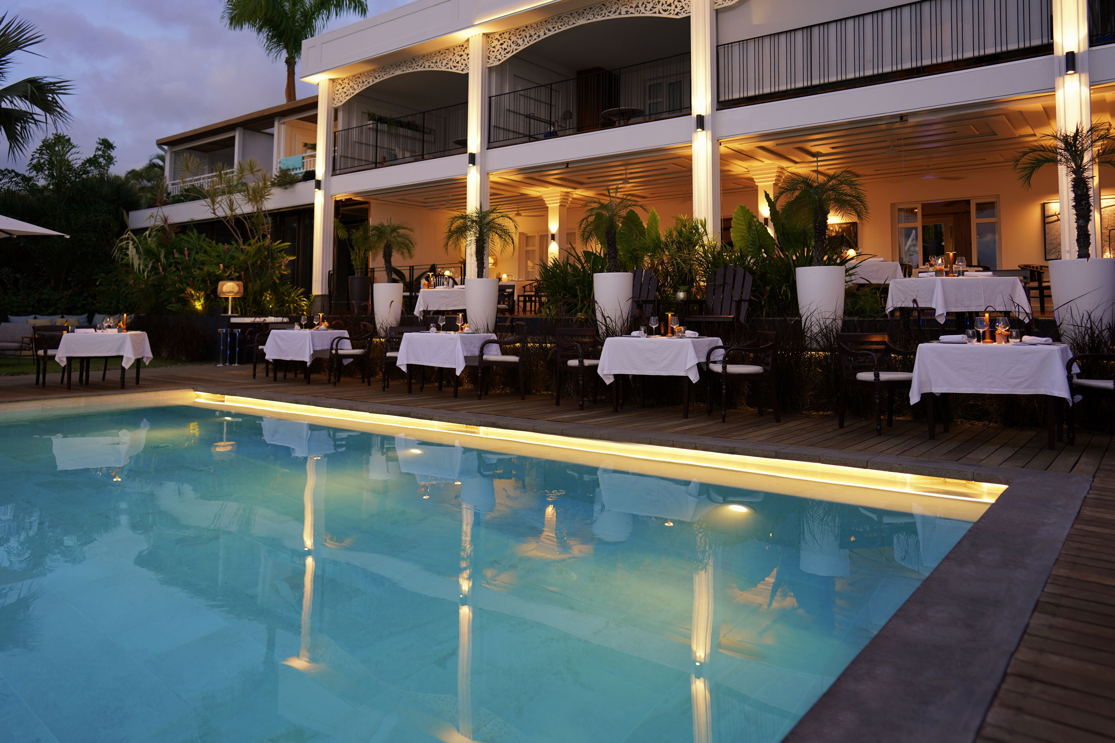 A poolside dining area with tables set for two, surrounded by palm trees and a pool.