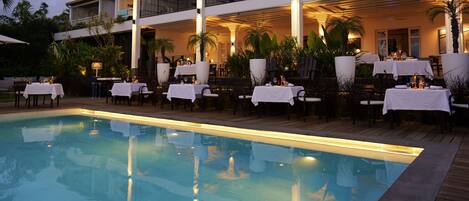 A poolside dining area with tables set for two, surrounded by palm trees and a pool.