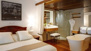 Une chambre d’hôtel comprenant un lit, un coin salon avec un fauteuil, une coiffeuse avec un miroir, une douche et une salle de bain avec des serviettes.