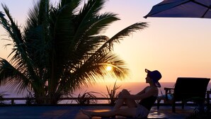 Une personne se détend au bord d’une piscine, avec une vue sur le coucher de soleil, un palmier et une chaise longue.