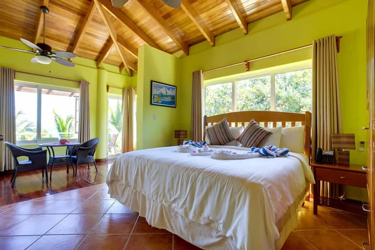 A hotel room with a large bed, a desk with chairs, a window with curtains, and a ceiling fan.