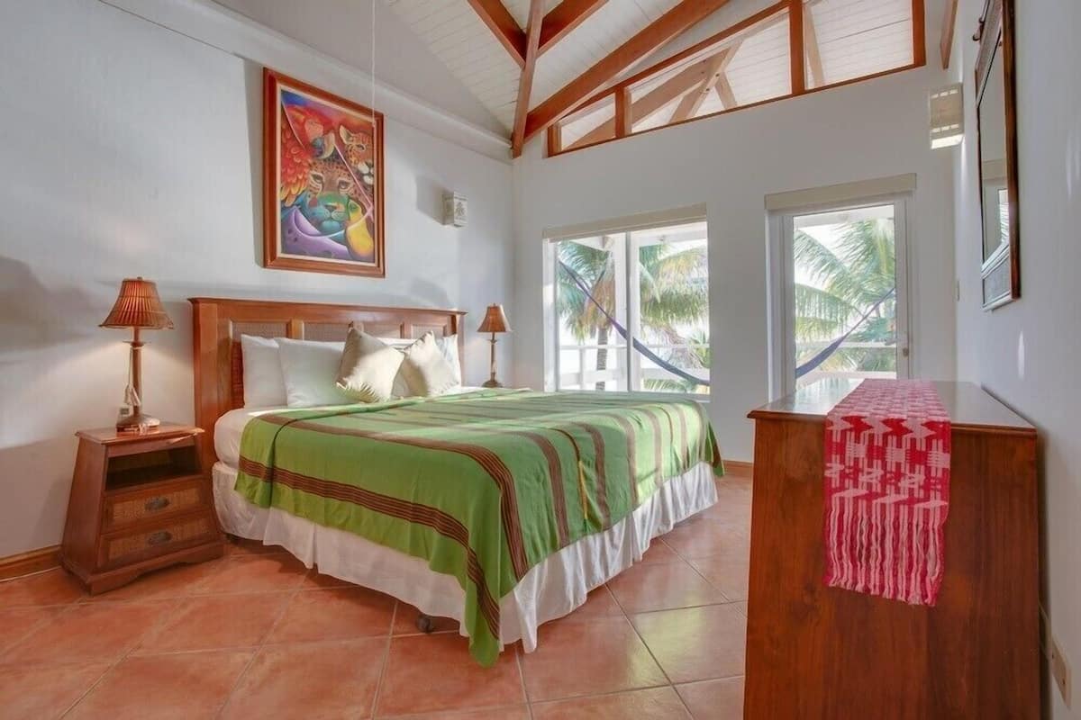 A bedroom with a bed, nightstands, a wooden headboard, a painting, and a window with a view of palm trees.