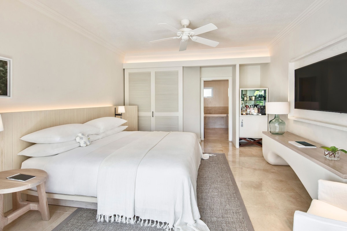 A hotel room with a bed, bedside tables, a TV, a ceiling fan, and a minibar.