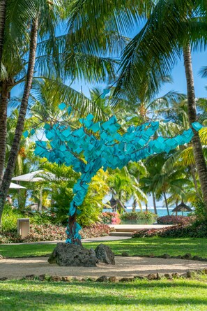 Una escultura de un árbol con hojas azules en un jardín tropical con palmeras y vista al océano.