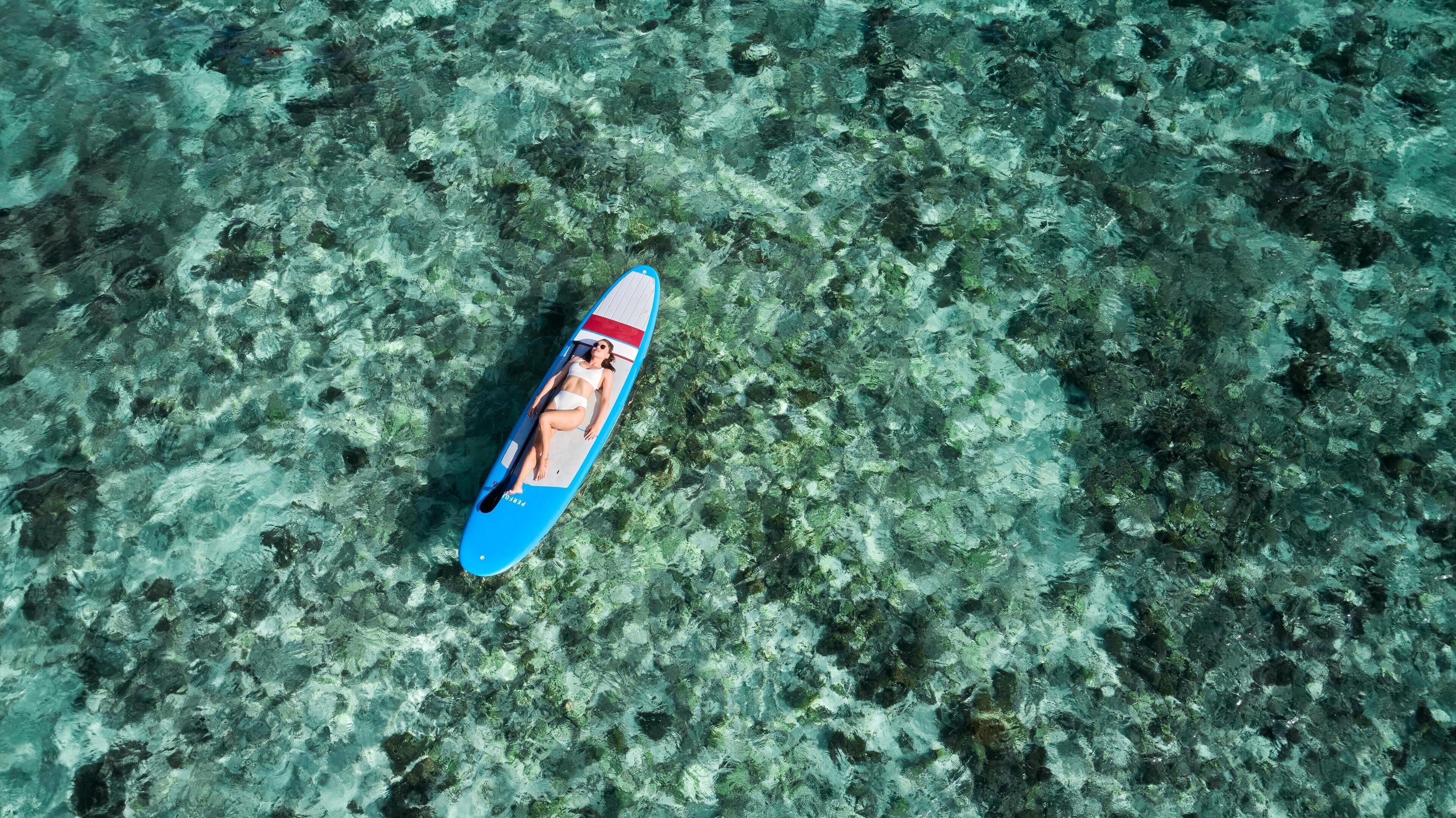 Vista aérea que muestra una tabla de remo azul con una persona acostada, rodeada de un entorno submarino poco profundo y rocoso.