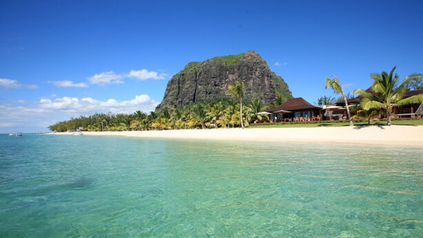 Una playa con agua turquesa cristalina, una gran montaña al fondo y un resort con palmeras y un edificio de techo de paja.