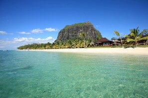 A beach with clear turquoise water, a large mountain in the background, and a resort with palm trees and a thatched-roof building. - LUX* Le Morne (Le Morne)