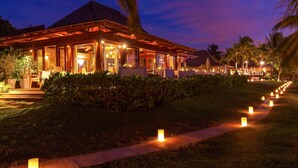 Un resort con un área exterior cubierta, un camino bordeado de velas y palmeras al fondo.