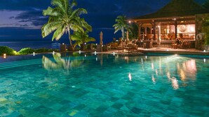 Vista nocturna de una piscina en un resort con agua cristalina, palmeras y un edificio con techo de paja.