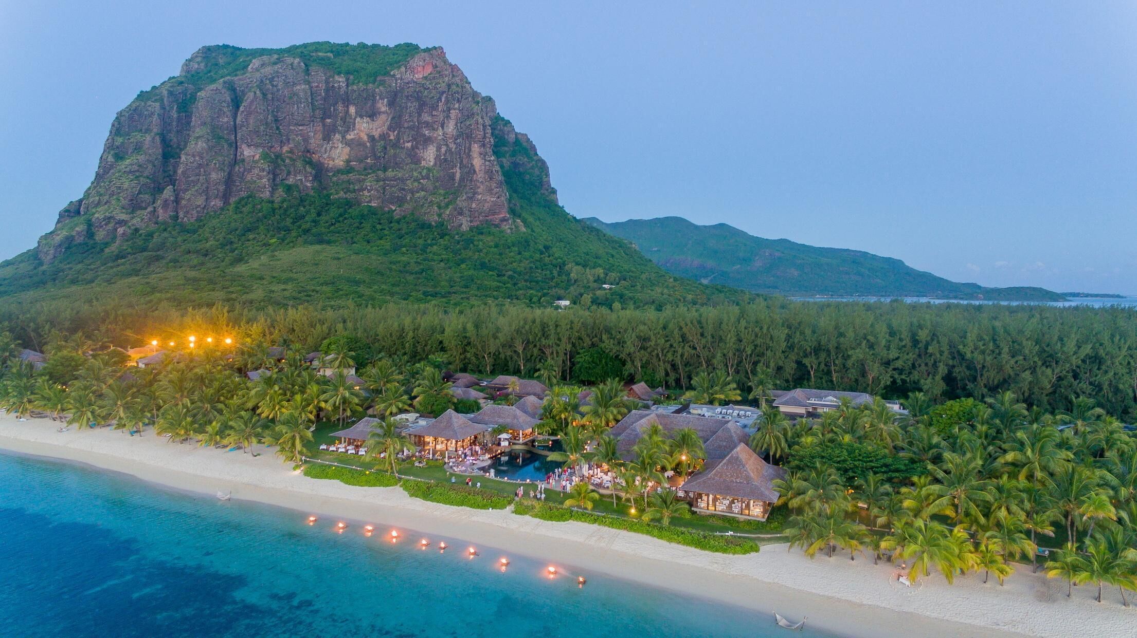A resort with thatched-roof buildings, a beach, and a mountain in the background.