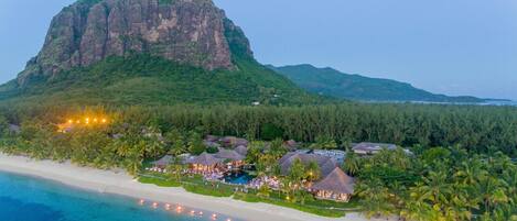 A resort with thatched-roof buildings, a beach, and a mountain in the background.
