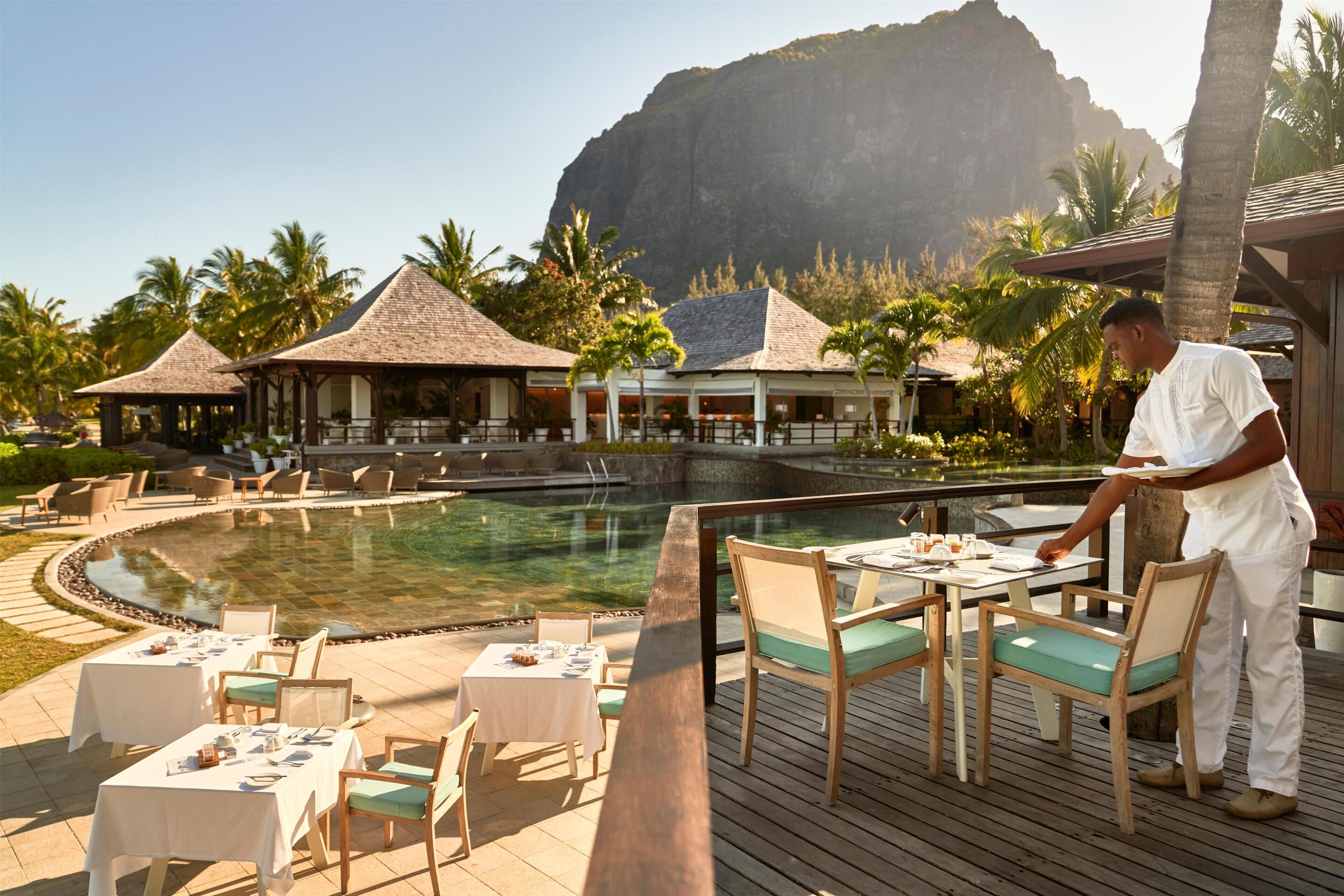 A poolside dining area with tables and chairs, a waiter, and a mountain backdrop.