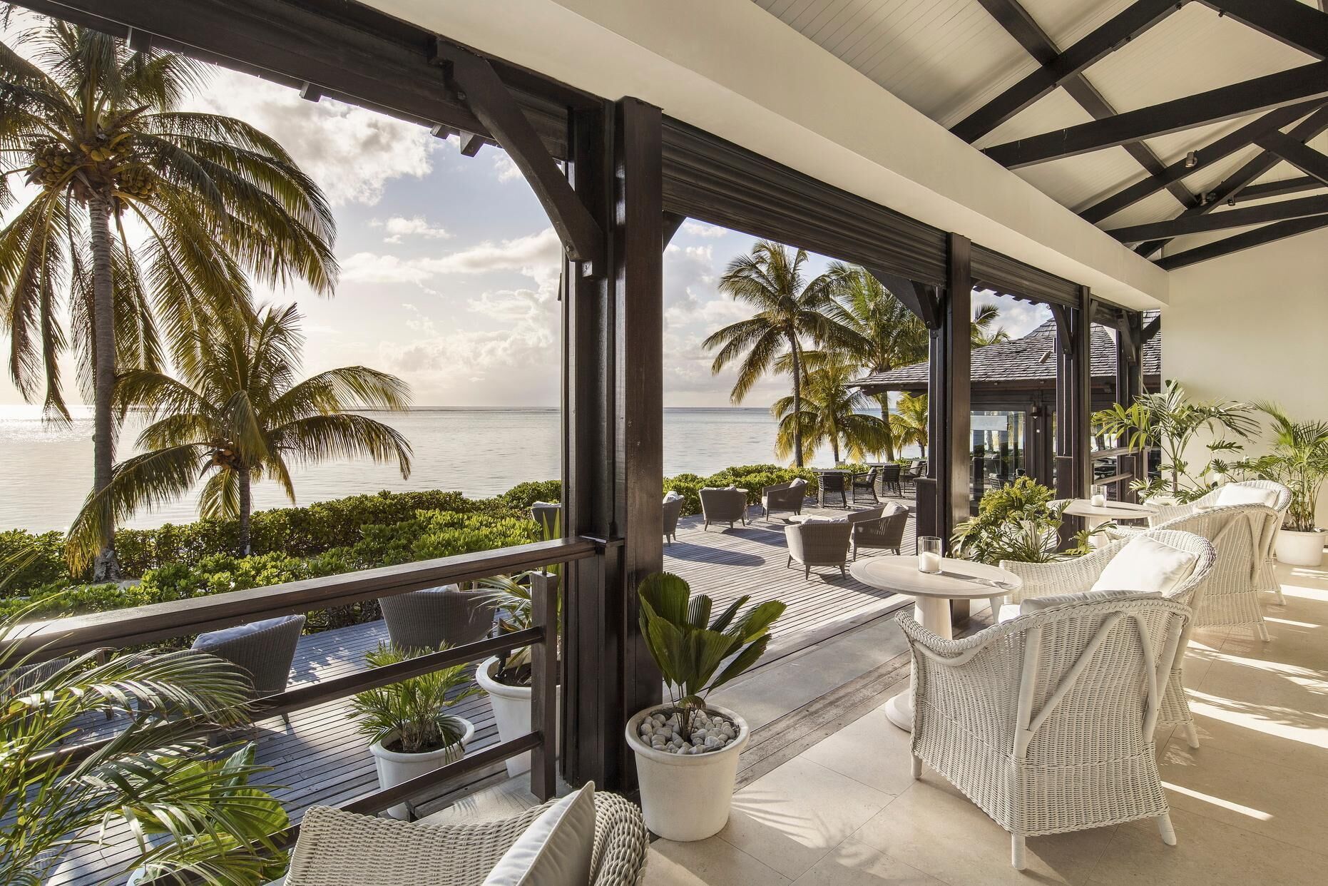 A covered outdoor seating area with wicker furniture, potted plants, and a view of the ocean.