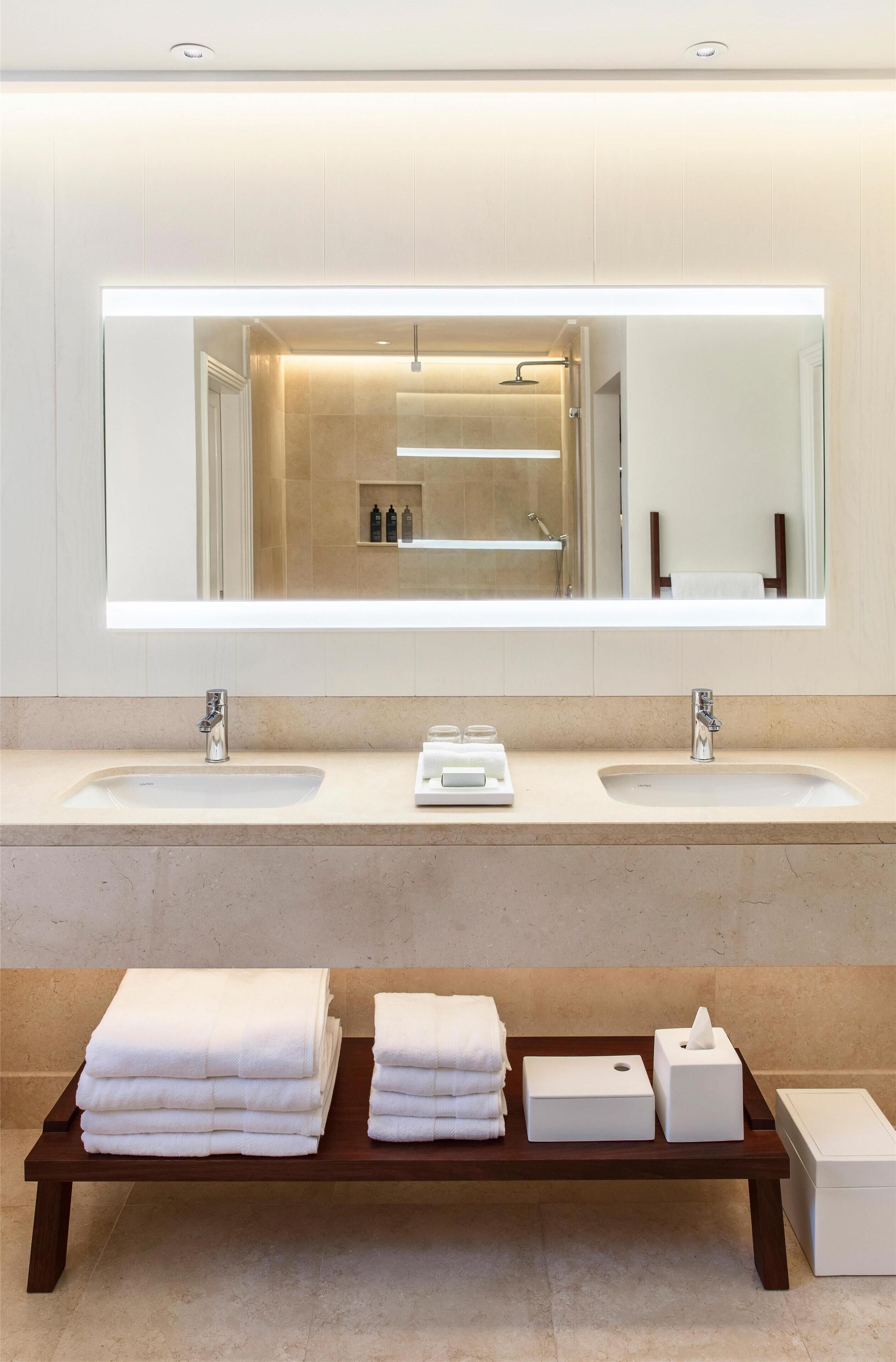 Un baño moderno con dos lavamanos, un espejo grande y un banco de madera con toallas y artículos de aseo.