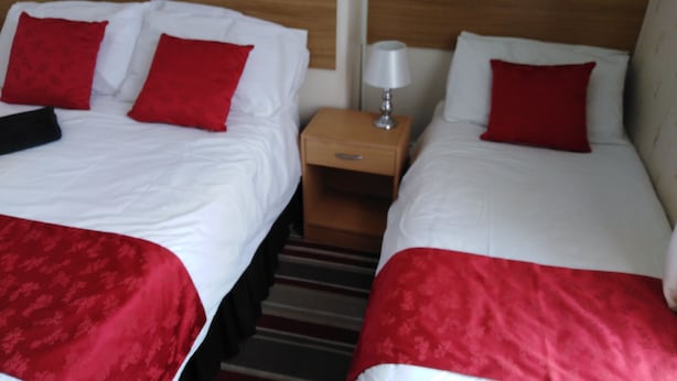 Dos camas con ropa de cama roja y blanca, un cabecero de madera, una mesita de noche con lámpara y una alfombra a rayas.