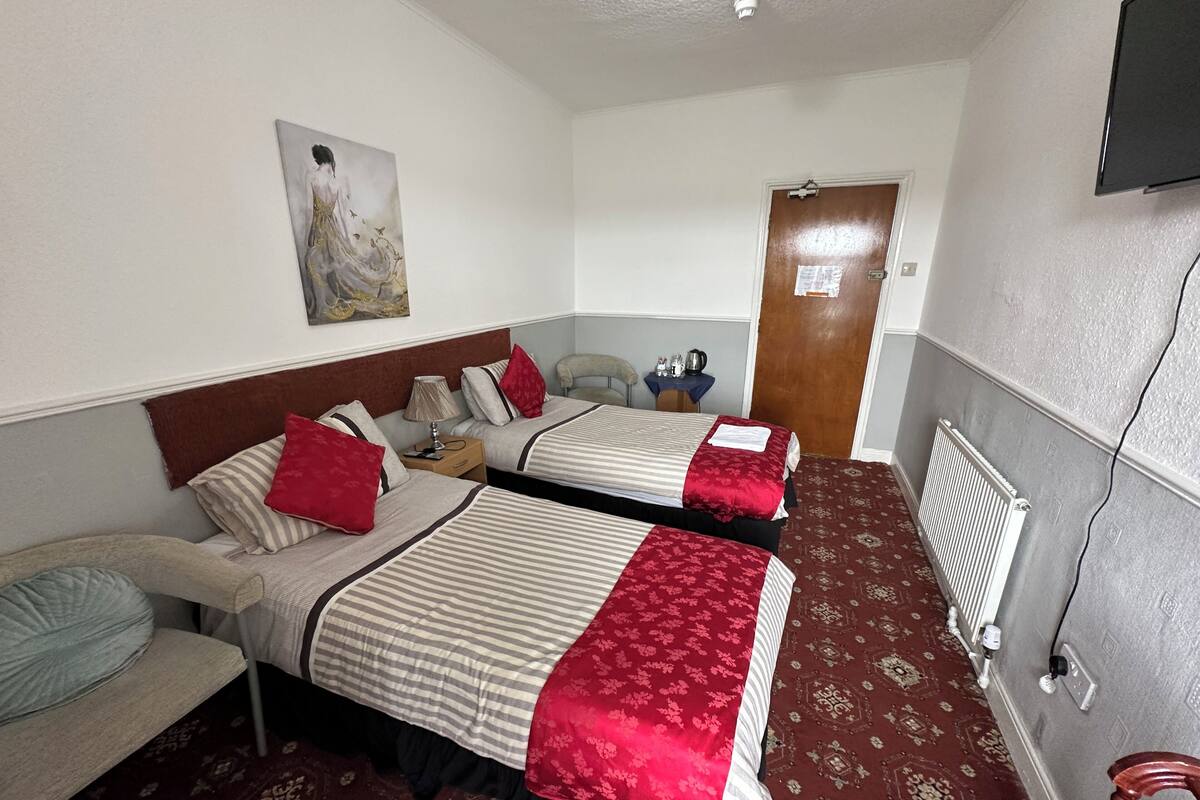 A hotel room with two beds, a chair, a table with a kettle, a painting on the wall, and a door.