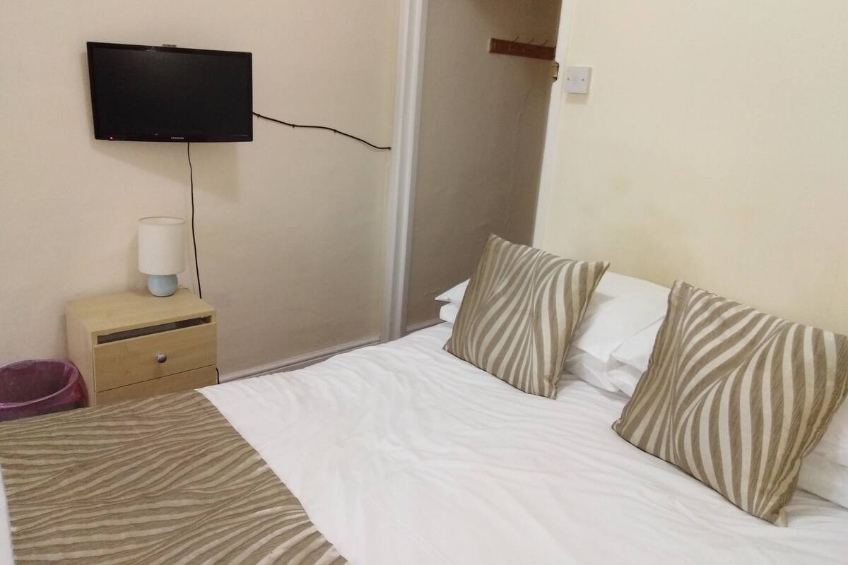 A hotel room with a bed, a television mounted on the wall, a bedside table with a lamp, and a door leading to another room.