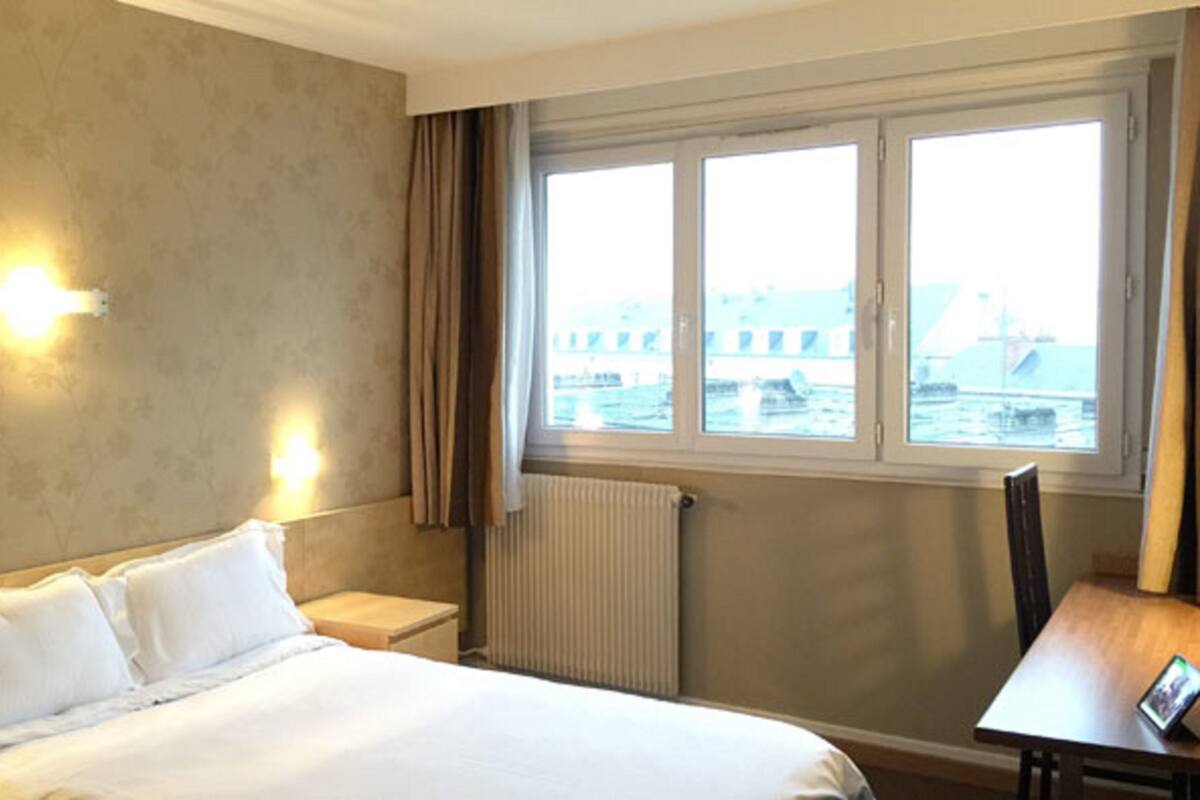 A hotel room with a bed, bedside table, lamp, and a view of a ship through the window.