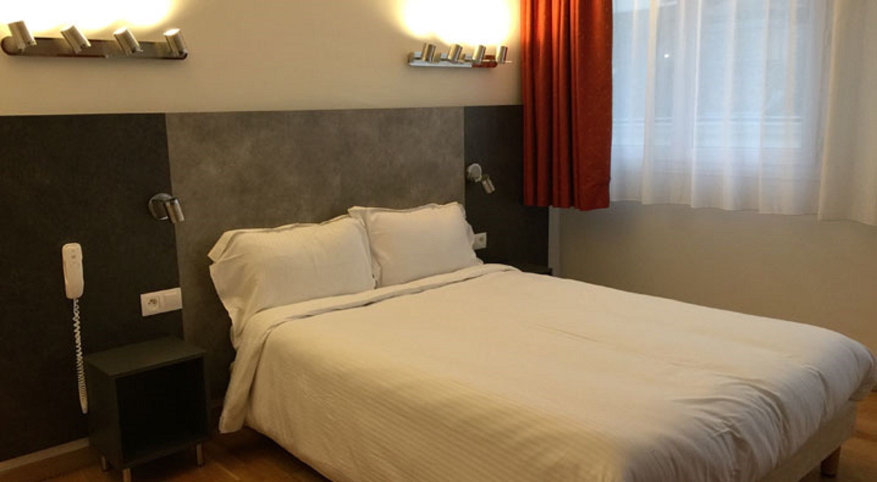 Una habitación de hotel con cama, mesitas de noche, teléfono y ventana con cortinas.