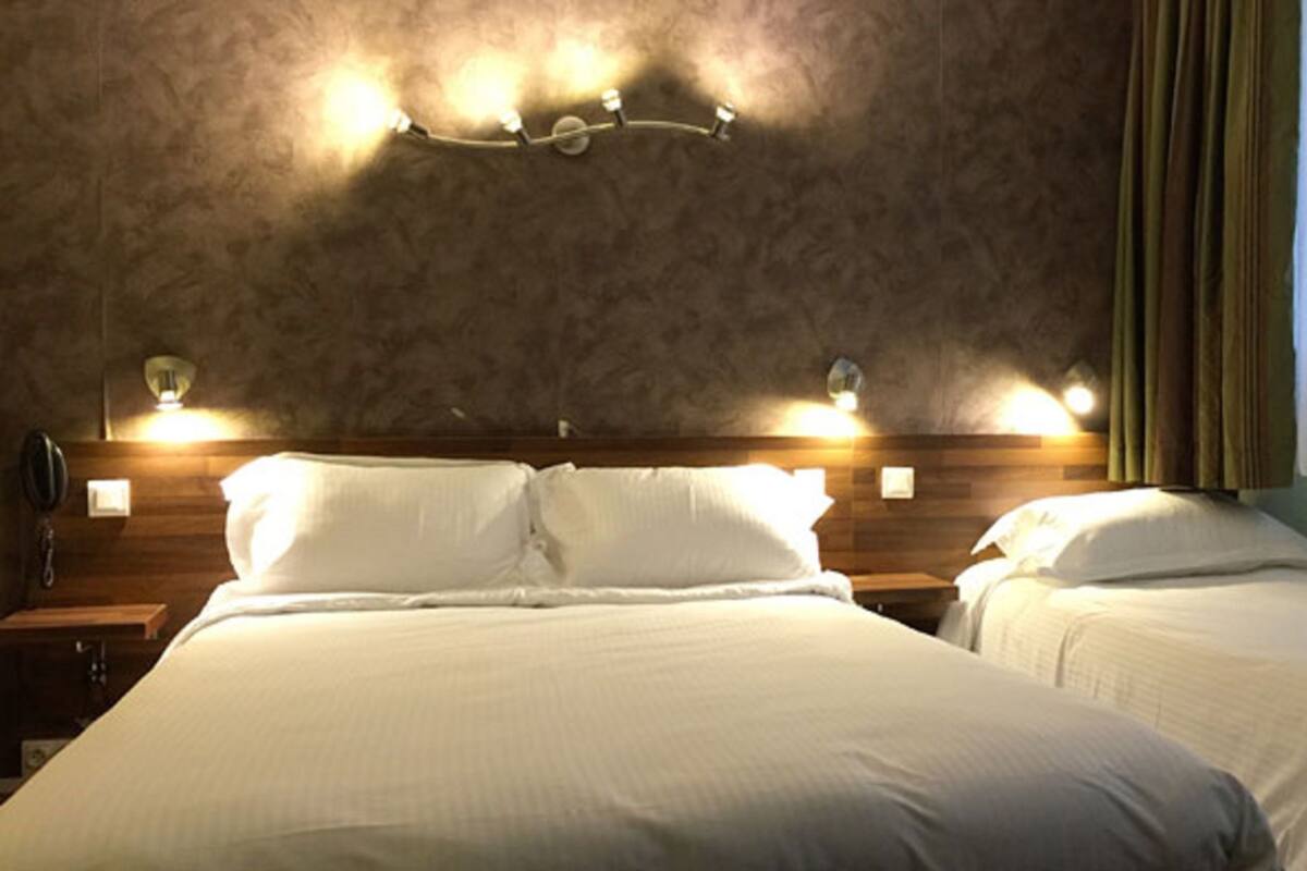 A hotel room with a large bed, two bedside tables, a wall-mounted light fixture, and a window with curtains.