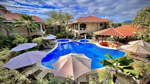 A pool area with multiple lounge chairs and umbrellas, surrounded by lush greenery and a building with a red-tiled roof. - Monte Carlo Luxury Condominiums (Jacó)