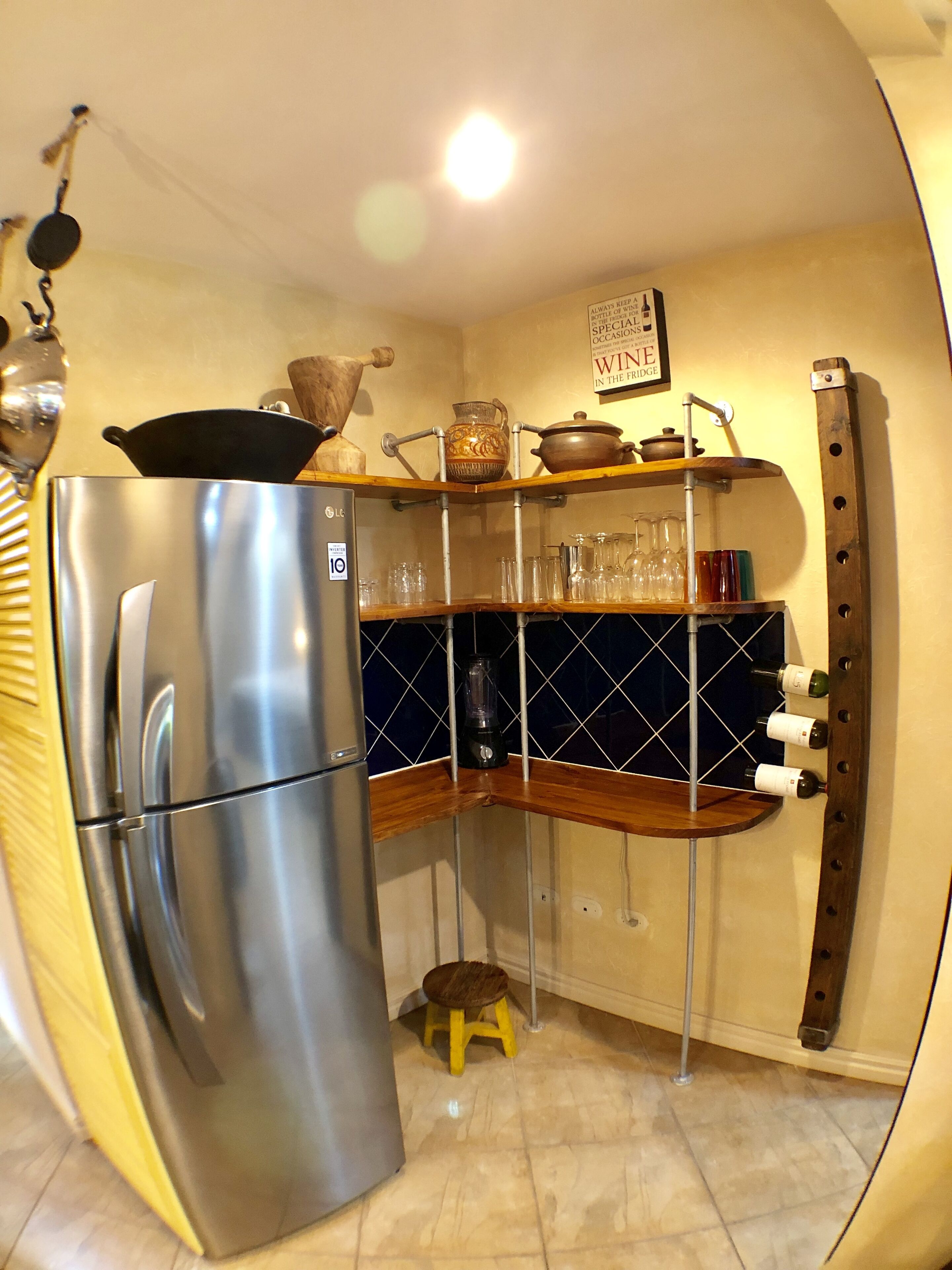 Una cocina compacta con un refrigerador de acero inoxidable, un taburete de madera y una estantería para vinos junto a la pared.