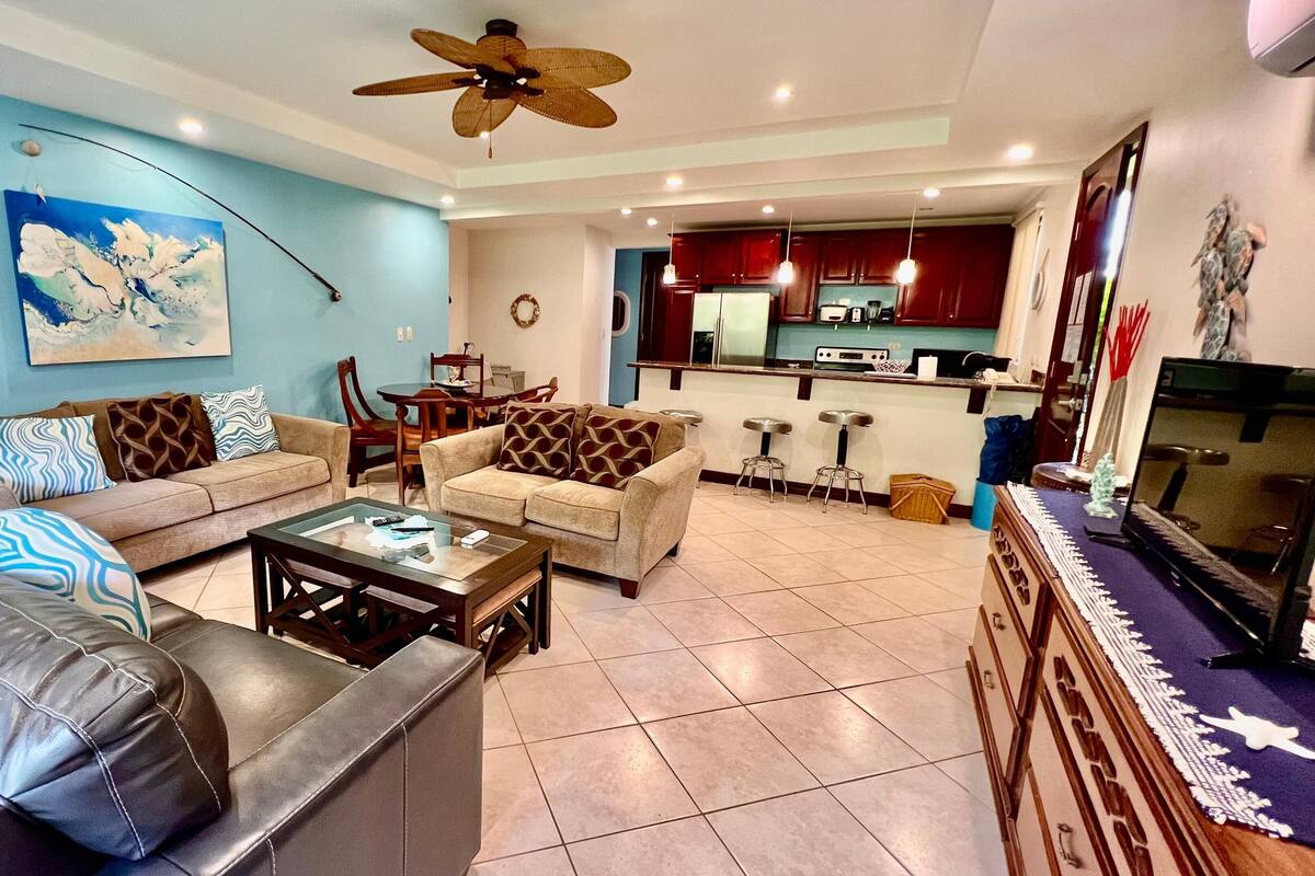 A living room with a sofa, a coffee table, and a TV. There is a kitchen in the background with a refrigerator and a microwave.