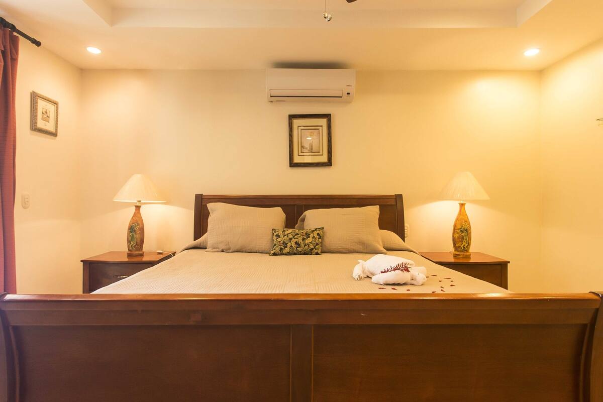 A neatly made bed with pillows and a blanket, two bedside tables with lamps, an air conditioner, and a framed picture on the wall.