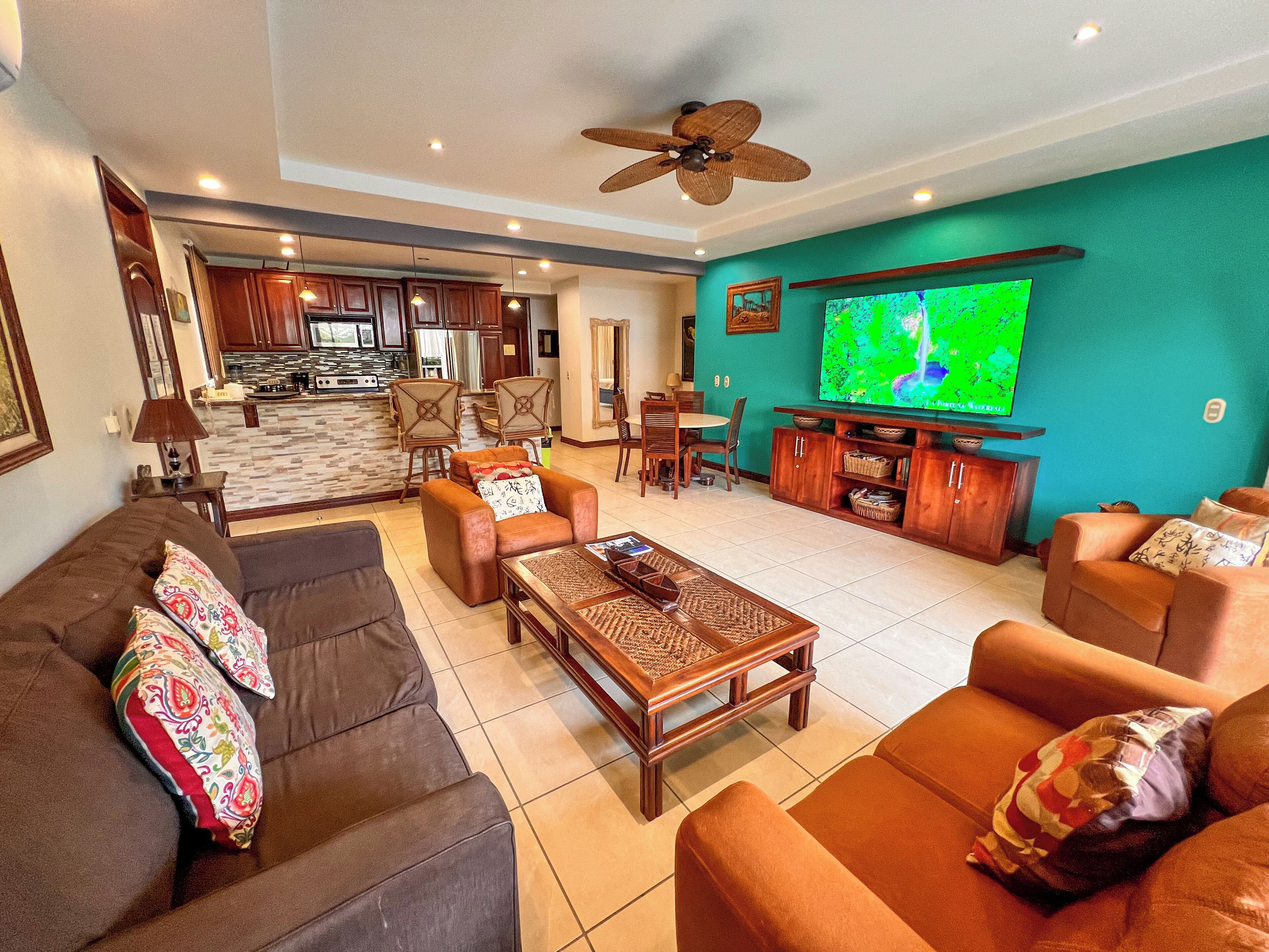 A spacious living room with a large TV, comfortable seating, a dining area, and a kitchen in the background.